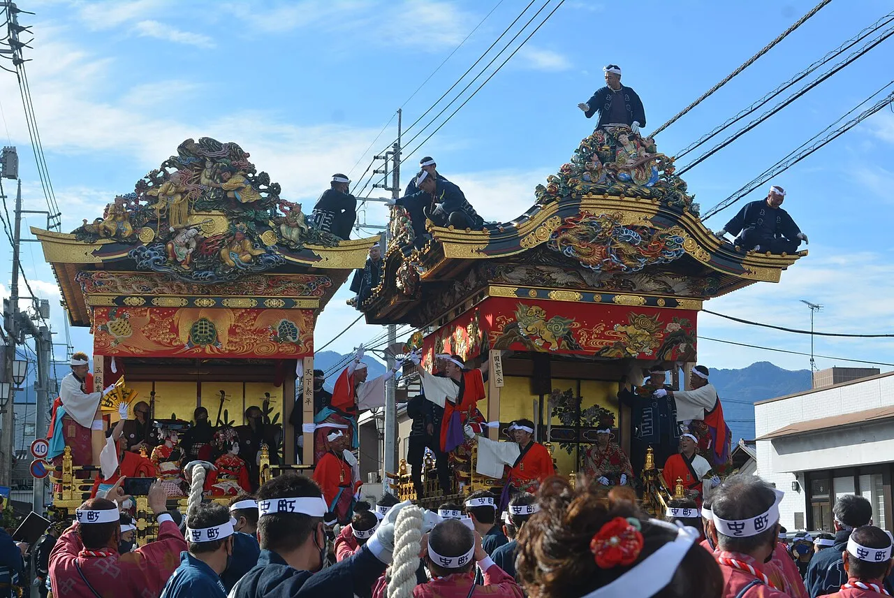 The Nakamachi and Kamimachi yatai floats, carved and lacquered, with crew members riding on their roofs above a crowd in festival coats.