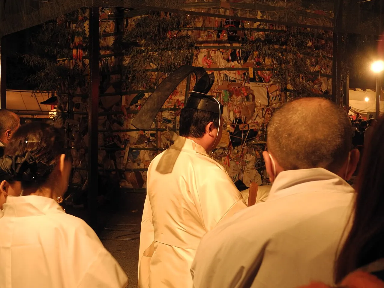 A Shinto priest in white robes and a black lacquered cap stands before a tall frame packed with old charms and talismans, ready to be burned.