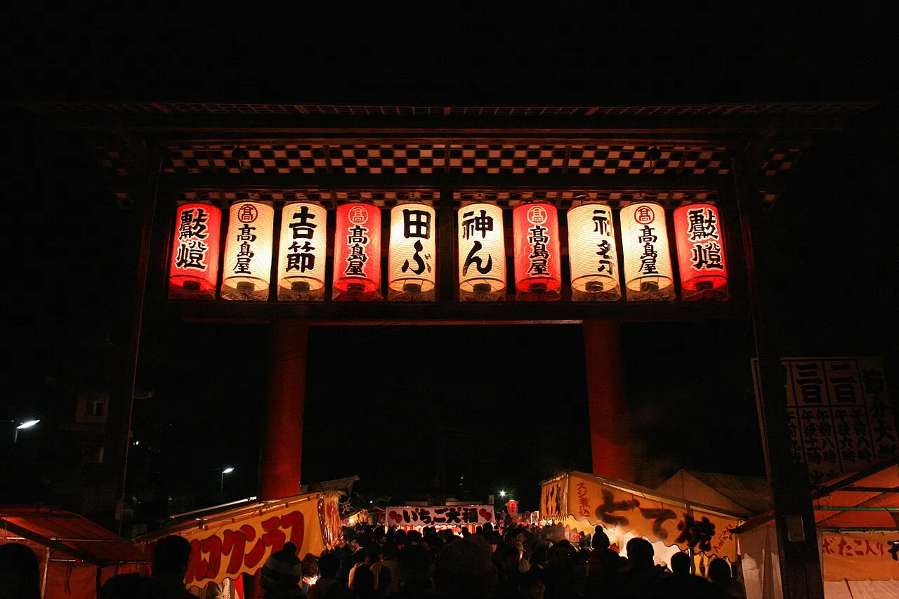 A large shrine gate hung with glowing red and white lanterns at night, above a crowd and rows of food stalls.
